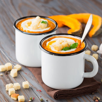 Homemade pumpkin soup with cream, croutons and basil in enamel mugs on rustic wooden background, selective focus