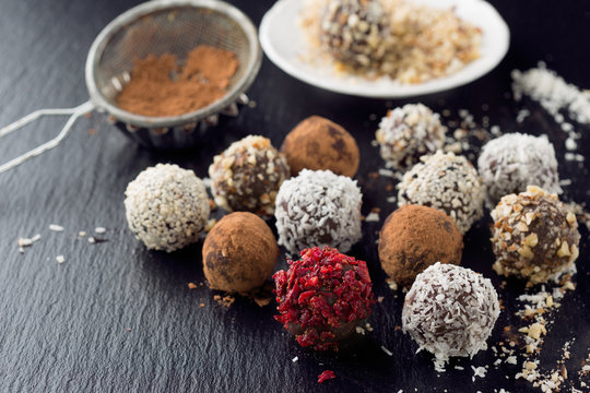 Homemade chocolate and nuts candy balls with cocoa powder, coconut, berries and chopped hazelnuts on black stone background, selective focus