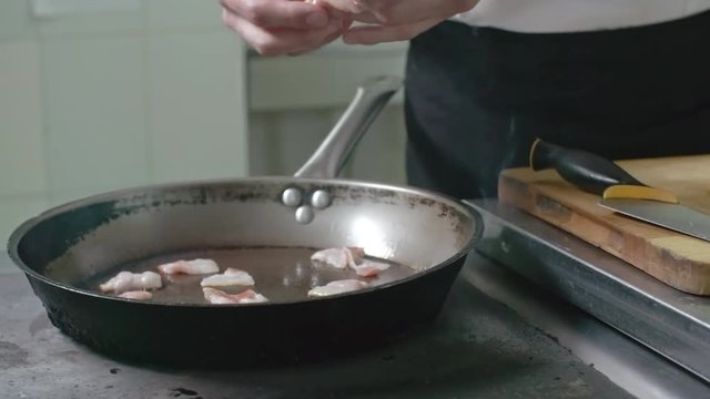 Closeup of hands of chef placing bacon strips on hot frying pan in the restaurant kitchen