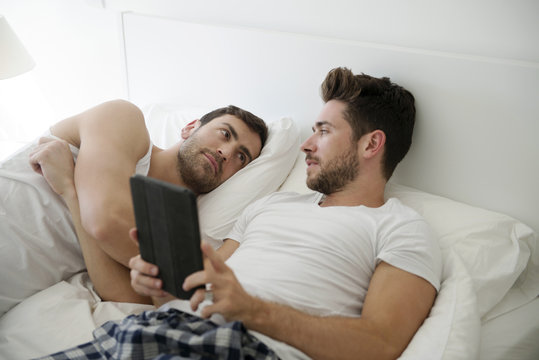 Gay Male Couple In Bed At Home Looking At Digital Tablet