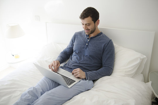 Man Relaxing In Bed At Home Looking At Laptop Computer