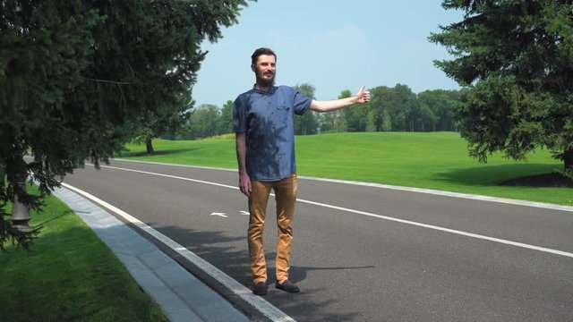 Young man traveling hitchhiking. Hitchhiker use hand signal point thumb up. Handsome guy stand near the road and waiting for some car. Caucasian traveler wearing in casual dress on the route.