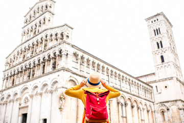 Young female traveler in yellow sweater and hat standing in front of San Michele basilica in Lucca old town in Italy. Having great vacations in Lucca. Back view - obrazy, fototapety, plakaty