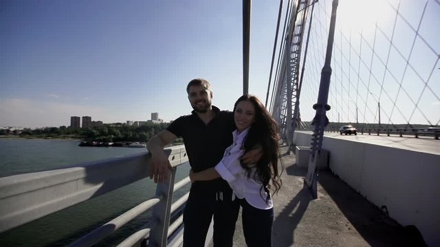 European girl hugging guy and lays down her head on his shoulder slo-mo. Man and woman looking at the camera in slow motion. Beautiful caucasian couple of people standing on the bridge slow-mo hd.