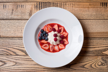 White milky creamy pudding with jam and berries blueberries, strawberry, raspberry on a white plate at the wooden background. Close-up top view