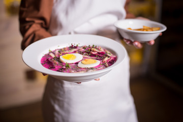 Plates with cold beetroot soup with cabbage, egg and with fried potatoes on a white plates in hand of woman with white apron. Close-up