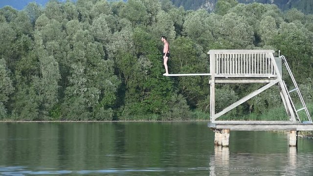 Man jumping from springboard into the lake reflecting beautiful landscape of green trees, slow motion