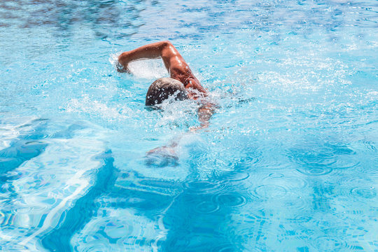 Senior man swimming front crawl, freestyle in an outside pool on a summer, sunny day. The pool steps can be seen and the splashes from the swim.