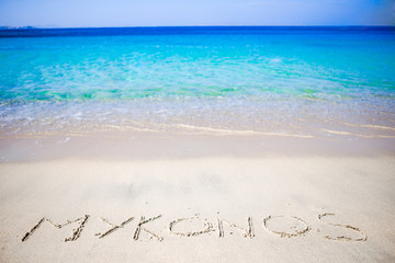 Word Mykonos handwritten on sandy beach with soft ocean wave on background - obrazy, fototapety, plakaty