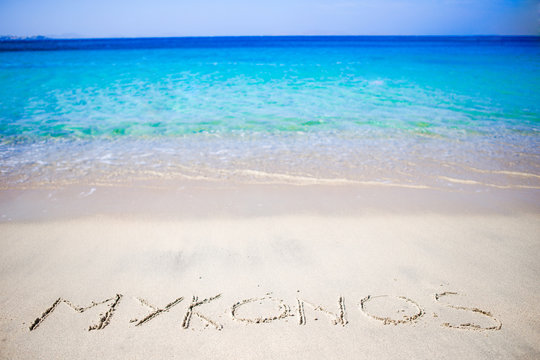 Word Mykonos handwritten on sandy beach with soft ocean wave on background