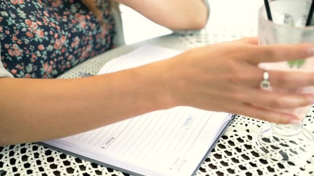 Girl writing something in her calendar and drinking cocktail at the table
