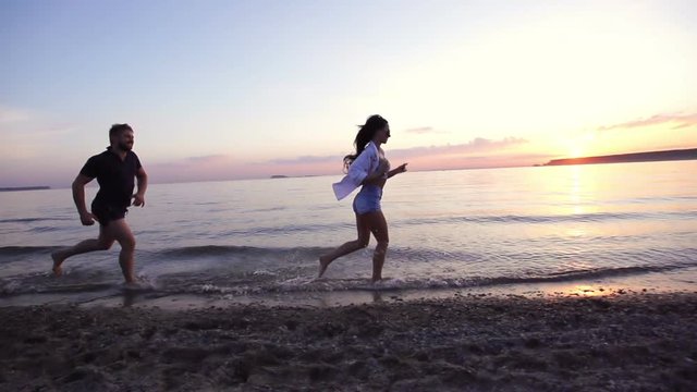 Couple people running in shallow water at sunset in slow motion. Spray from the fleeing feet Man and girl on waterfront sea in slow-mo. Crazy jogging on the beach 20s. Jog boyfriend and girlfriend.