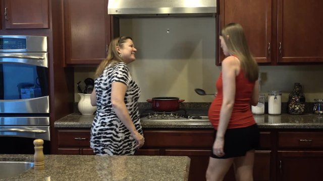 A mother and her expecting daughter prepare food in the kitchen.  	