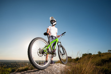 Fototapeta na wymiar Athlete guy in helmet and glasses standing with the mountain bicycle on the hill under blue sky. Bottom view