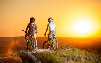 Cyclists in helmets and glasses stay on the mountain bikes at the precipice of slope against evening sky with bright sun and looking at the sunset. Blurred background