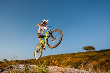 Naklejka na ściany i meble Male biker making dangerous jump on a mountain bike on the slope against blue sky. Cyclist is wearing sportswear helmet and glasses. Bottom view