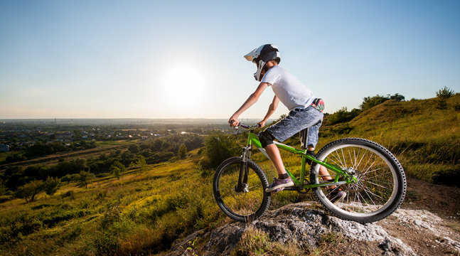 Biker in helmet and glasses on mountain bike stands on the precipice of hill and looking down under blue sky against greenery and small town into the distance. Wide angle view