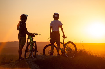 Silhouette of young bikers staying with mountain bicycles on the top of mountain against evening sky with bright sun and looking at the sunset