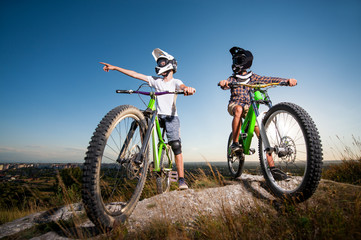 Two bicyclists wearing in helmets and glasses standing with the mountain bicycles on the hill under blue sky. Men look away and show up on something. Wide angle view