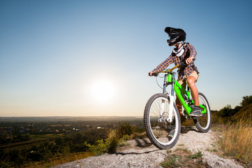 Bicyclist in helmet and glasses on mountain bike stands on the precipice of hill under blue sky against greenery and small town in the distance. Bottom view.