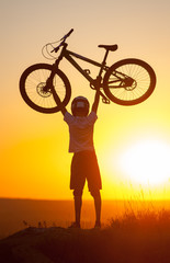 Silhouette of young cyclist in helmet holding a mountain bike over himself on the top of hill against evening sky with bright sun at the sunset