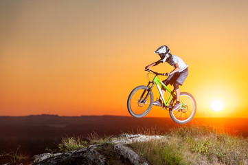 Fototapeta na wymiar Sunset. Male biker riding downhill on a mountain bike on the precipice of hill against evening sky with bright sun. Cyclist is wearing sportswear helmet and glasses. Bottom view