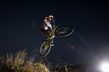 Obraz premium Bicyclist making high jump on a mountain bike on the hill against night sky. Cyclist is wearing sportswear helmet and glasses. Bottom view. Extreme downhill
