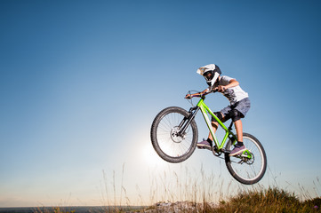 Fototapeta na wymiar Young biker riding downhill and making dangerous jump on a mountain bike from the slope against blue sky and bright sun. Cyclist is wearing white sportswear helmet and glasses.