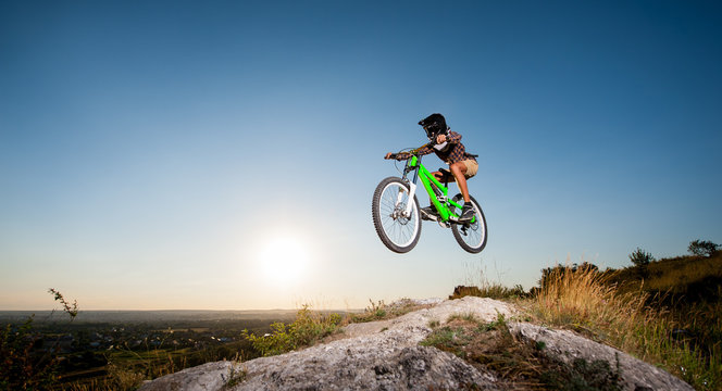Young bicyclist in helmet and glasses making high jump on a mountain bike on the hill against blue sky. Bottom view