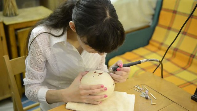 Young adult woman artisan is starting to carve the big white ostrich eggshell by high-speed rotation tool for high-precision work to make openwork cross-cutting shell carving in the home room.