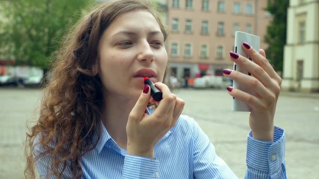 Pretty girl using smartphone as a mirror while painting her lips
