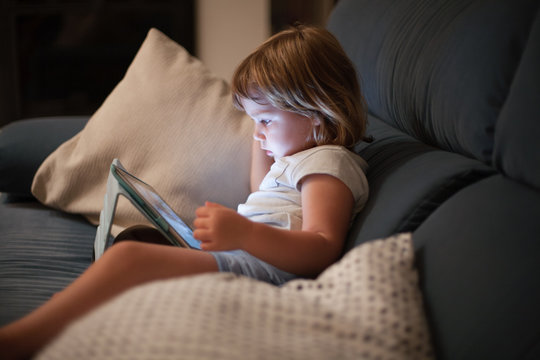 Blonde Three Years Old Baby Shirt And Shorts, Sitting Comfortably In Sofa Inside Home At Night Reading And Watching Digital Tablet, Face Illuminated By The Light Of The Screen
