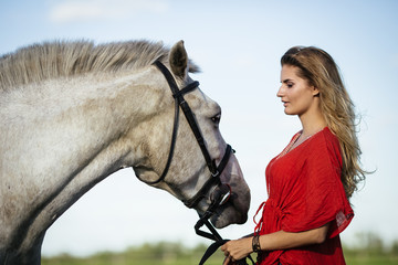Obraz premium Portrait of young beautiful woman dressed in red tunic with horse in the meadow
