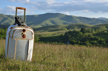Suitcase with binoculars and sunglasses on mountain meadow with hills and sky in background