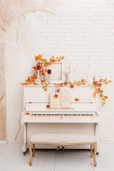 White piano decorated with yellow leaves in front of old white brick wall. Autumn season in photo studio.