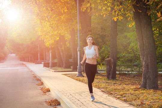 young beautiful happy and sexy runner girl with nice body in the autumnal colored park. with sun on backlight is looking at camera with happiness and smile with white sport suit.