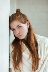 Closeup portrait of young adorable redhead woman wrapped in a white knitted blanket posing near white wall indoor natural light