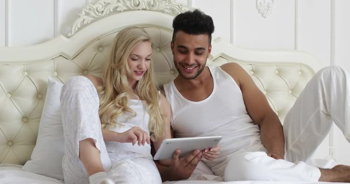 Couple lying bed using tablet computer, mix race man woman smile morning bedroom