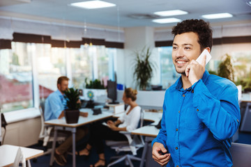 Young successful businessman speaking on phone, smiling, over office background.