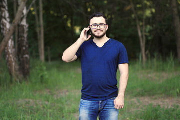 Man is talking on the phone. Hi is wearing denim pants, blue t-shirt and glasses. He has got black hair, brown eyes and white skin 