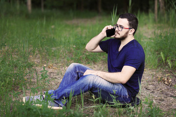 Sitting man is talking on the phone. Hi is wearing denim pants, blue t-shirt and glasses. He has got black hair, brown eyes and white skin 