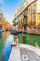 Fototapeta na wymiar Woman sits near a canal and admires gondolas and architecture of Venice. Italy.