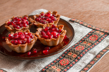 Four tartletes with custard and pomegranate in brown plate on linen tablecloth with ethnic pattern.