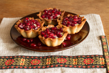 Four tartletes with custard and pomegranate in brown plate on linen tablecloth with ethnic pattern.