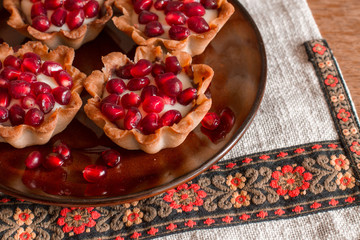 Four tartletes with custard and pomegranate in brown plate on linen tablecloth with ethnic pattern.