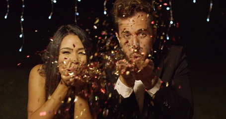Young couple celebrating New Year blowing colorful paper confetti over their hands towards the camera against twinkling party lights