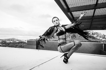 Black and white photo of beautiful seductive angel woman wearing lingerie and leather belts crouching on the roof over cloudy sky