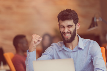 Young bearded caucasian modern business man sitting in a startup office using laptop, looking downward the screen, smiling - business, work, technology concept