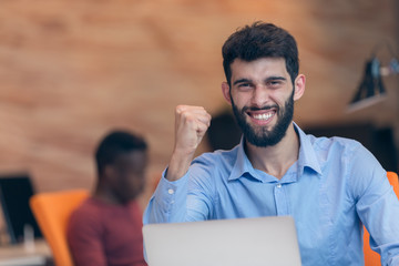 Young bearded caucasian modern business man sitting in a startup office using laptop, looking downward the screen, smiling - business, work, technology concept