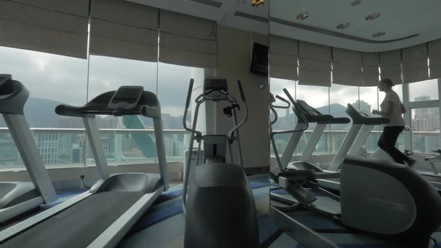 Steadicam shot of young woman running on treadmill in the gym and looking to Hong Kong through panoramic window of upper floor